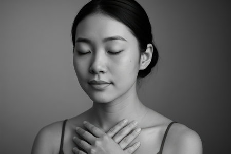 Calm young woman meditating with closed eyes and hands on chest in serene monochrome indoor setting, practicing mindfulness.の素材