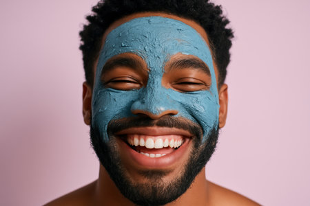 Smiling man with a blue facial mask enjoying skincare on a pastel background, showing healthy complexion and self care routine.の素材