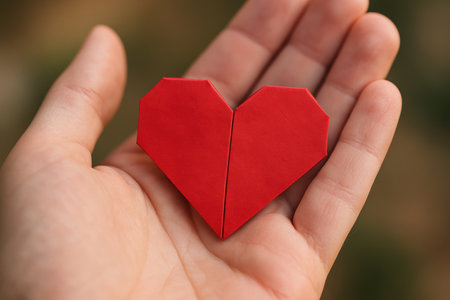 Close up of a hand gently holding a red origami heart symbolizing love, care, and thoughtful handmade craftsmanship outdoors.の素材