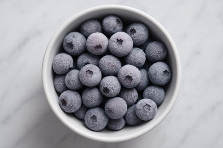 Frozen blueberries in white bowl on marble surface, overhead view showing frosty texture and healthy snack option.の素材