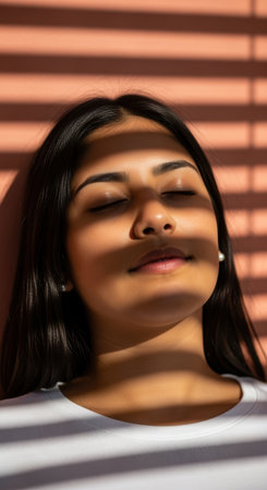 Calm young woman relaxing with eyes closed as sunlight and window blinds create striped shadows on her face indoors.の素材