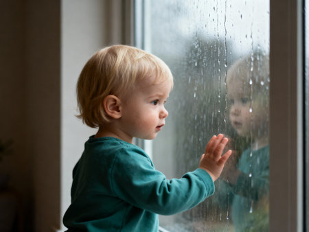 Curious young child gazing out rain covered window, touching glass while watching droplets on a gloomy day indoors.の素材