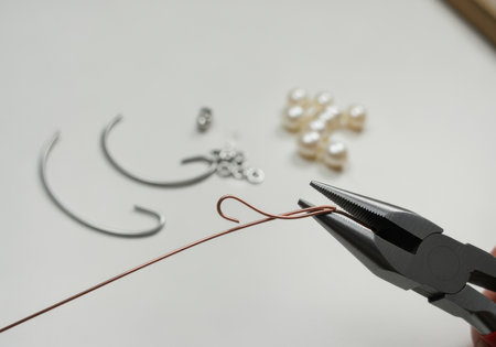 Hand crafting delicate wire jewelry with pliers, assembling metal components and pearls on a clean white workspace for handmade design.の素材