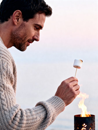 Man roasting marshmallow over open flame outdoors in cozy sweater during dusk by tranquil lakeside.の素材