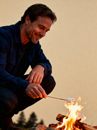 Smiling man roasting marshmallow over campfire at dusk, enjoying outdoor adventure and relaxation in nature.の素材