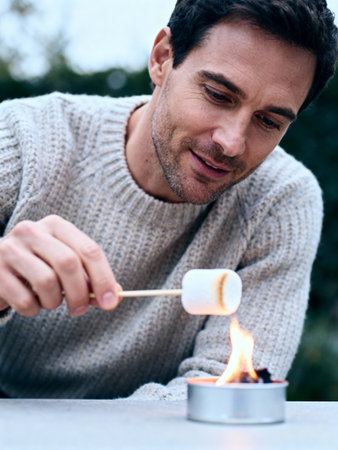 Man in cozy sweater roasting marshmallow over small flame outdoors, enjoying a relaxing autumn evening in natural surroundings.の素材