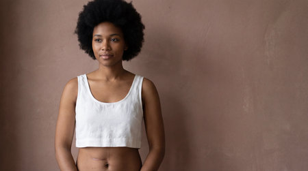 Confident young woman with natural afro hairstyle standing in minimalist indoor setting, wearing white crop top against neutral wall.の素材