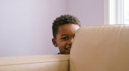 Smiling young boy playfully peeking from behind beige sofa in bright living room with soft natural light and minimal interior decor.の素材