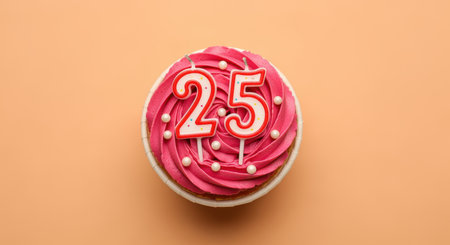 Pink frosted cupcake with number 25 candles and white candy pearls on pastel peach background for birthday celebration.の素材