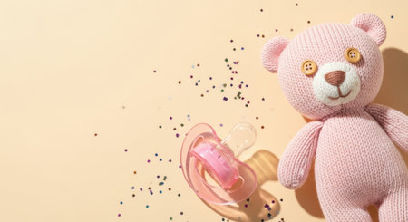 Soft pink knitted plush bear and pacifier on pastel background with colorful confetti for newborn baby girl celebration concept.の素材