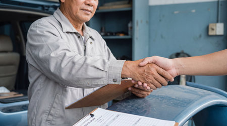 Mechanic shaking hands with customer in auto repair shop after completing car service and signing maintenance agreement on clipboard.の素材