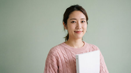Young woman in pink sweater smiling confidently while holding a white binder against a soft green studio background.の素材