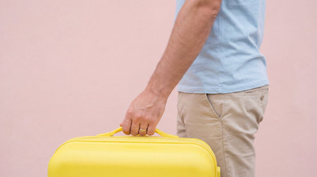 Man holding bright yellow suitcase against pastel pink wall, cropped torso view emphasizing casual summer outfit and travel concept.の素材