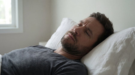 Middle aged man lying in bed with tired expression in soft morning light beside window and plain white wall interior.の素材