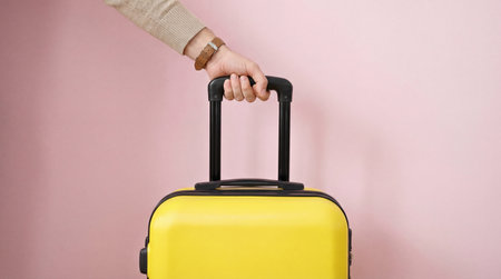 Hand holding yellow suitcase against pastel pink background symbolizing travel preparation and modern minimalist vacation concept.の素材