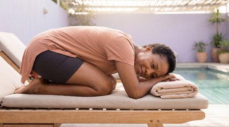 Relaxed woman resting in child's pose on poolside lounge chair, smiling with head on folded towel in peaceful outdoor wellness setting.の素材