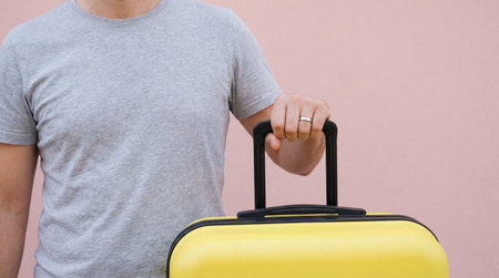 Man holding yellow suitcase against pastel pink wall, cropped torso view of casual traveler standing with hand on luggage handle.の素材
