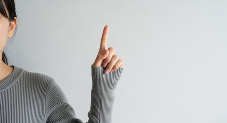 Woman raising index finger against light grey background in casual sweater, making gesture for attention, instruction or counting one.の素材