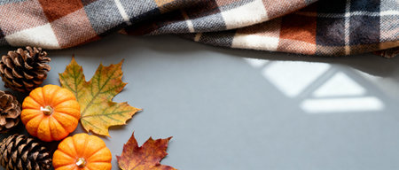 Cozy autumn scene with orange pumpkins, pine cones, and colorful leaves next to a warm plaid blanket on a gray background.の素材