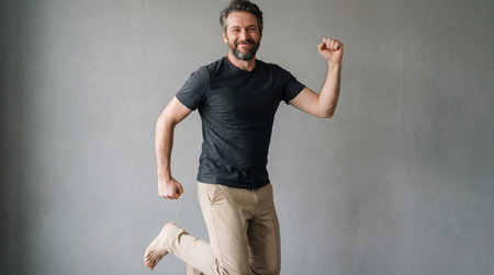 Happy barefoot middle aged man joyfully jumping indoors against plain grey wall in casual t shirt and beige pants.の素材