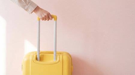 Hand holding handle of yellow suitcase against soft pastel wall, minimalist travel concept with light and shadow details.の素材