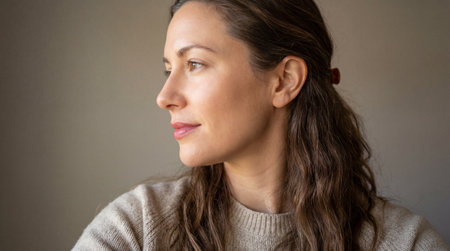 Calm young woman gazing sideways in soft natural light with relaxed expression and casual sweater against neutral indoor background.の素材