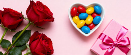 Red roses, pastel heart bowl with colorful candies, and a pink gift box tied with satin ribbon on a soft pink background.の素材