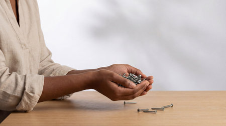 Person holding assorted metal screws in hands above wooden table surface with scattered hardware in soft natural light.の素材