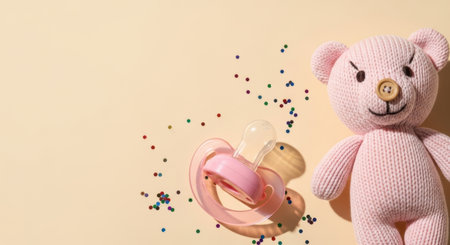 Soft pink knitted plush bear with baby pacifier and colorful confetti on pastel beige background for newborn celebration.の素材