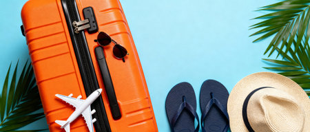 orange suitcase with sunglasses, airplane toy, flip flops, and straw hat arranged on blue background with tropical palm leaves.の素材