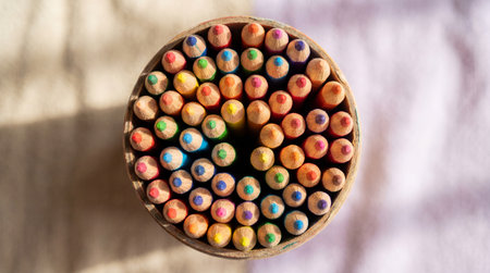 Top view of colorful wooden pencils arranged in a circular pattern inside a cup on a softly lit neutral background.の素材