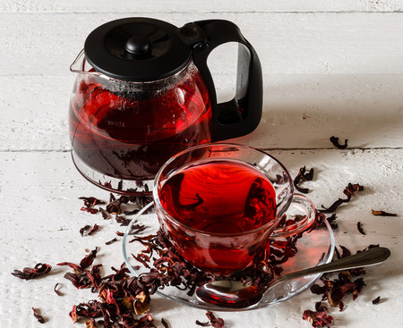 Cup of Karkadeh Red Tea with Dry Flowers and kettle on a white wooden tableの写真素材