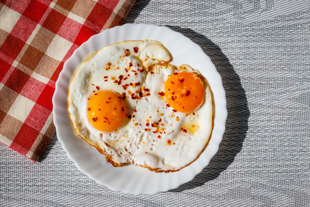 Fried eggs on a plate with chili peper spice on a table with red tablecloth.の写真素材