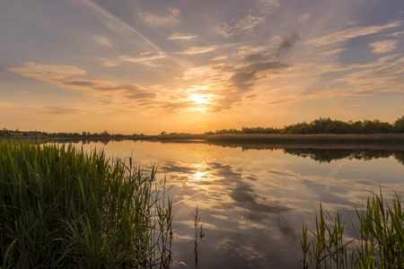 Scenic view of beautiful sunset or sunrise above the pond or lake at spring or early summer evening with cloudy sky background and reed grass at foreground. Landscape. Water reflection.の写真素材