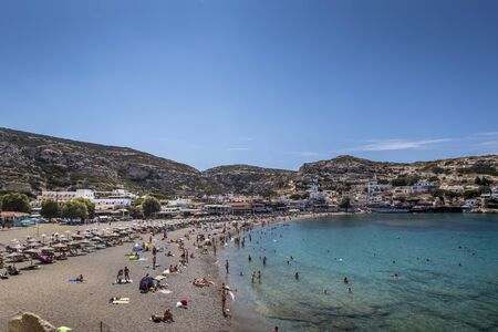 The sea coast and mountains of Matala on a clear summer sunny day with and a blue sky in the background. Crete, Greece. Sunbeds on the beach with tourists resting.の写真素材