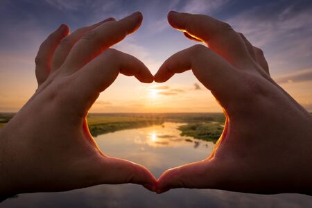 Hands in the shape of heart against the sunset over the lake or river in the summer with a cloudy sky background. Landscape.の写真素材