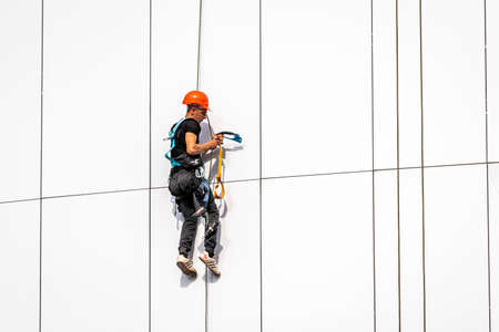 Russia, Kaluga - SEPTEMBER 16, 2020: An industrial climber hanging from a rope with a carabiner and securing the white cladding panels of a building at a construction site on a clear sunny day.のeditorial素材