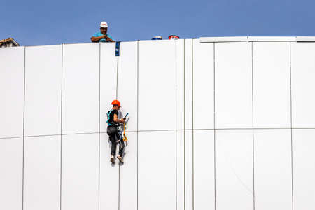 Russia, Kaluga - SEPTEMBER 16, 2020: An industrial climber hanging from a rope with a carabiner and securing the white cladding panels of a building at a construction site on a clear sunny day.のeditorial素材