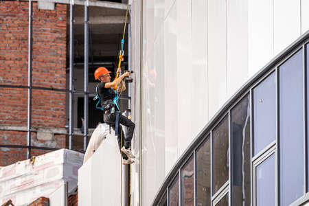 Russia, Kaluga - SEPTEMBER 16, 2020: An industrial climber hanging from a rope with a carabiner and securing the white cladding panels of a building at a construction site on a clear sunny day.のeditorial素材