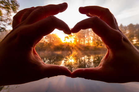 Male hands making a heart-shaped gesture against the background of sunrise or sunset on a river or...