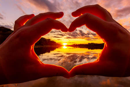 Male hands making a heart-shaped gesture against the background of sunset or dawn on a river or lake...