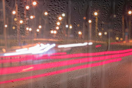 Defocused headlights from moving cars behind rainy, wet glass at night. Abstract concept of rainy...