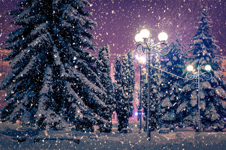 Winter night park with lanterns, pavement and trees covered with snow in heavy snowfall.
