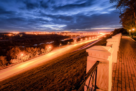 Moving car with blur light through city at night. Bridge over the river and the road. A view from the park from a height with a fence in the foreground. Cityscape.の写真素材