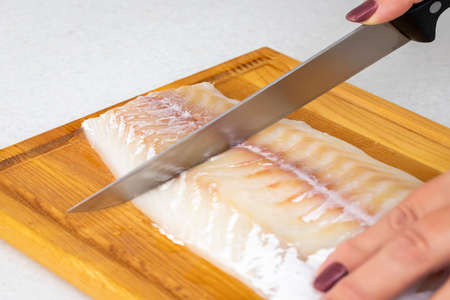 Female hands cutting white fish meat with a knife on a cutting board.の写真素材