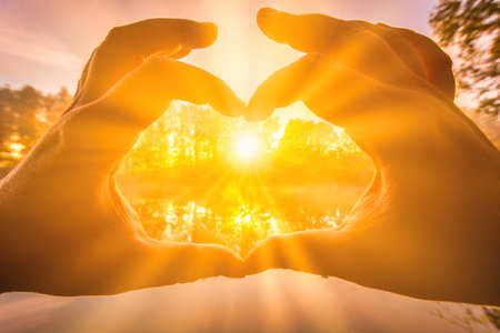 Male hands making a heart-shaped gesture against the background of sunrise or sunset on a river or lake in autumn morning.の写真素材