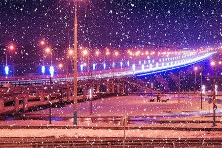Traces of headlights from cars moving at winter night on the bridge, illuminated by lanterns in a...