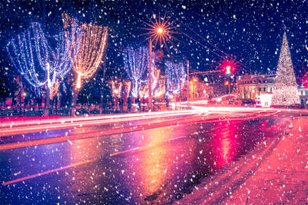 Night city with Christmas decorations, spruce and traces of headlights of moving cars, reflected in the wet asphalt in a snowfall.の写真素材