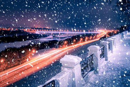 Snowfall in a winter park at night with lanterns, view to road with car motion, pavement and trees covered with snow.の写真素材