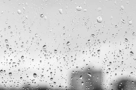Rain drops on window glasses surface with gray sky and buildinhs background. Natural backdrop of raindrops. The concept of bad rainy weather.の写真素材